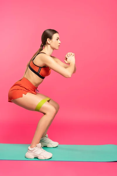 Side view of sportswoman with clenched hands doing squat with resistance band on pink — Stock Photo