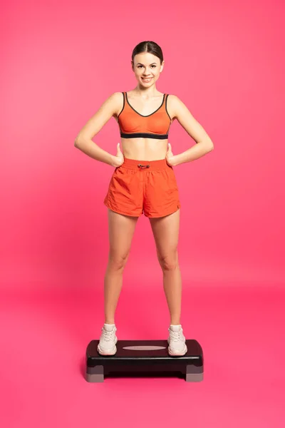 Happy sportswoman standing with hands on hips on step platform on pink — Stock Photo