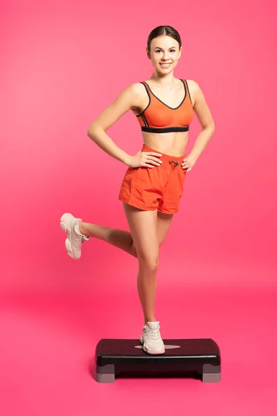 Happy sportswoman with hands on hips exercising on step platform on pink — Stock Photo