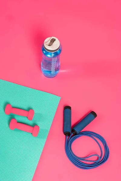 Vista superior de garrafa de esportes com água doce perto pulando corda e halteres no tapete de fitness em rosa — Fotografia de Stock