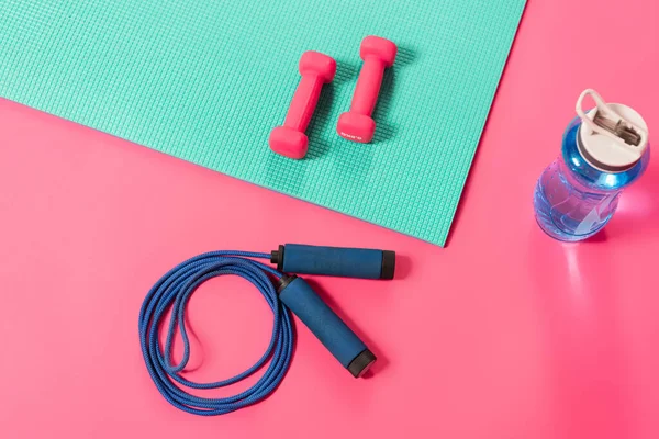 Top view of sports bottle with water near skipping rope and dumbbells on fitness mat on pink — Stock Photo