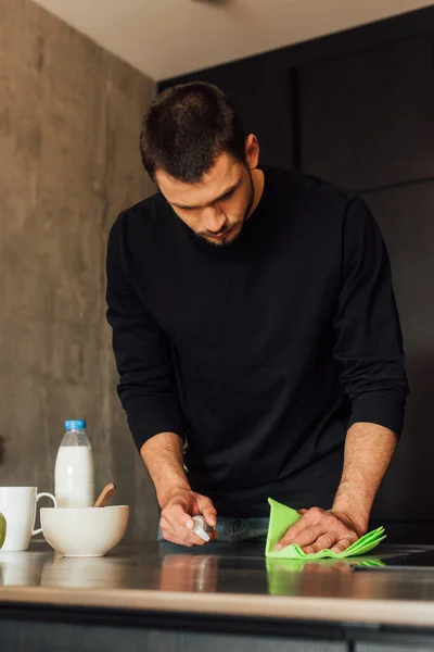 Homme barbu tenant bouteille avec liquide antibactérien et chiffon près de la table en bois — Photo de stock