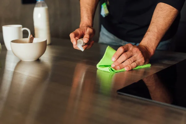 Vista recortada del hombre sosteniendo botella con líquido antibacteriano y trapo cerca de la mesa de madera - foto de stock