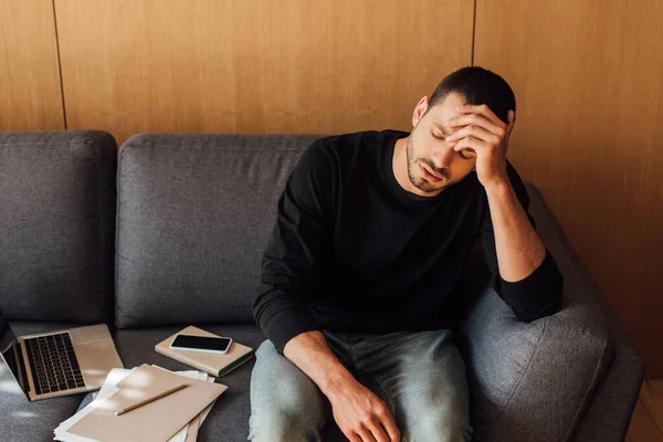 Sick man feeling unwell and touching head while sitting on sofa near laptop and smartphone with blank screen — Stock Photo