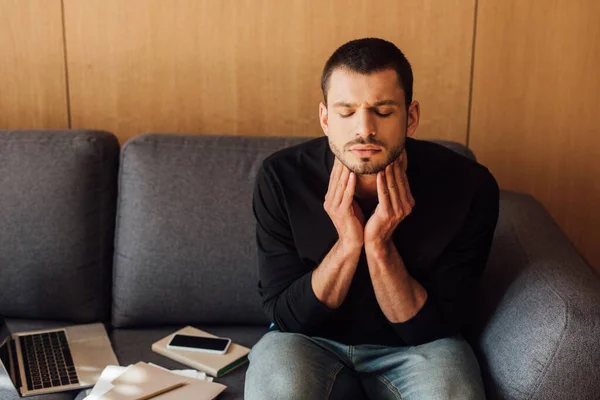 Kranker Mann mit geschlossenen Augen, der den Hals berührt, während er auf dem Sofa neben Laptop und Smartphone mit leerem Bildschirm sitzt — Stockfoto