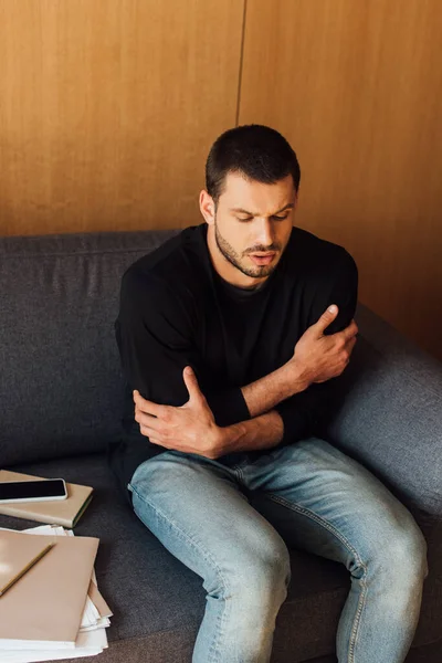 Sick man feeling cold while sitting on sofa near smartphone with blank screen — Stock Photo