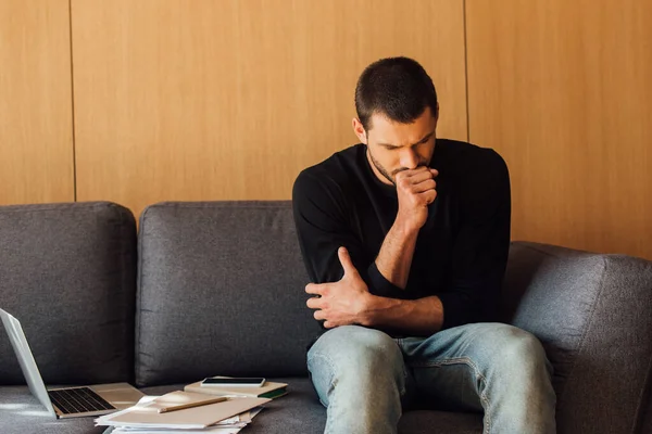 Hombre enfermo tosiendo mientras está sentado en el sofá cerca de la computadora portátil y teléfono inteligente con pantalla en blanco — Stock Photo