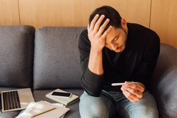 Sick and bearded man looking at digital thermometer near laptop and smartphone with blank screen — Stock Photo