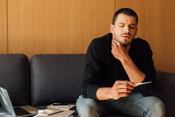 Sick man with sore throat looking at digital thermometer near laptop and smartphone with blank screen — Stock Photo