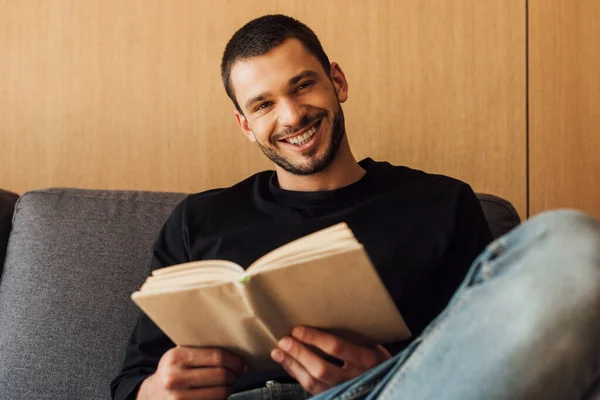 Fuoco selettivo di uomo allegro e barbuto che tiene libro in soggiorno — Foto stock