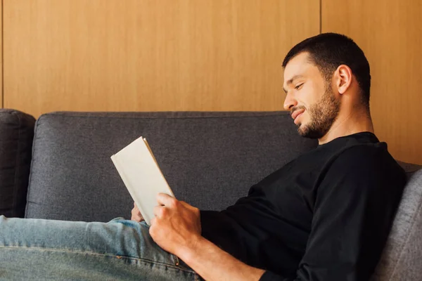 Vista lateral de homem bonito e barbudo leitura livro na sala de estar — Fotografia de Stock