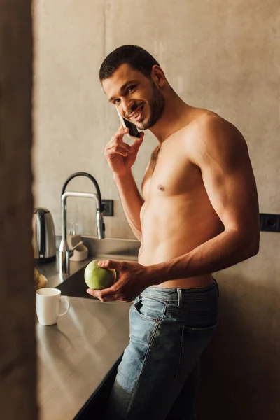 Foco seletivo de homem barbudo feliz segurando maçã e falando no smartphone perto da xícara na mesa na cozinha — Fotografia de Stock