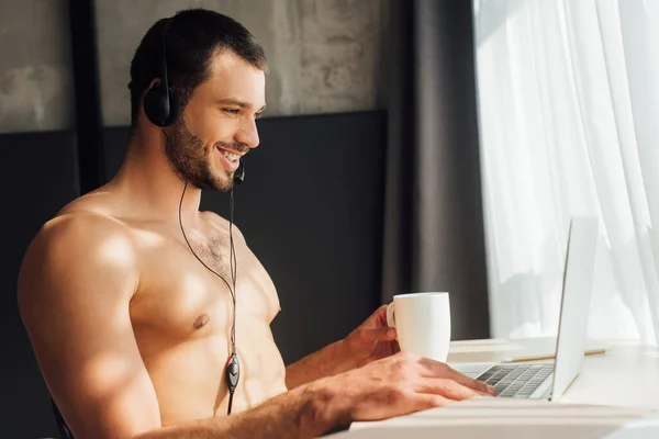 Operador feliz y sin camisa en la taza de la celebración de auriculares cerca del ordenador portátil en casa - foto de stock