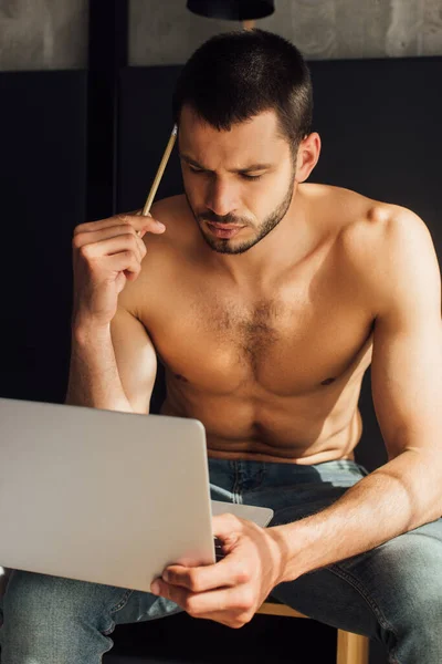 Красивый и без рубашки фрилансер держит карандаш и смотрит на ноутбук — стоковое фото
