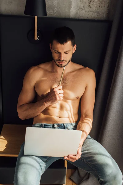 Barbudo y sin camisa freelancer sosteniendo lápiz y mirando a la computadora portátil - foto de stock