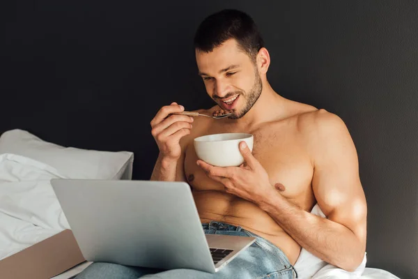 Homem feliz e sem camisa segurando tigela com colher e assistir filme no laptop — Fotografia de Stock