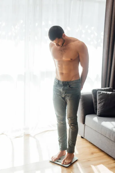 Muscular man standing on scales in living room — Stock Photo