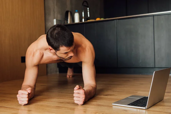 Бородатый и мускулистый человек делает упражнения доски во время просмотра онлайн тренировки на ноутбуке — стоковое фото