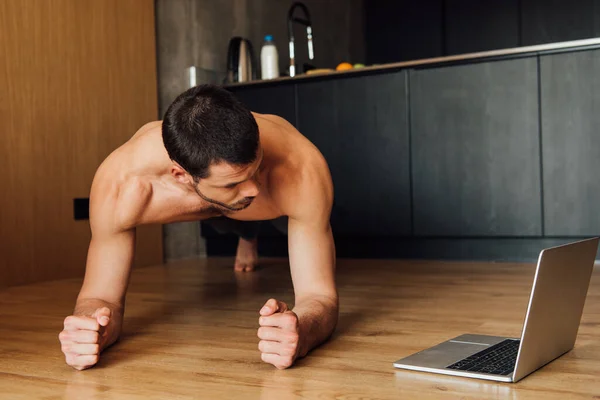 Hombre barbudo y musculoso haciendo ejercicio de tablón mientras mira el entrenamiento en línea en el ordenador portátil - foto de stock