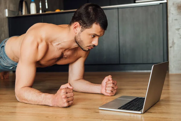 Homem muscular fazendo exercício prancha enquanto assiste treinamento on-line no laptop — Fotografia de Stock