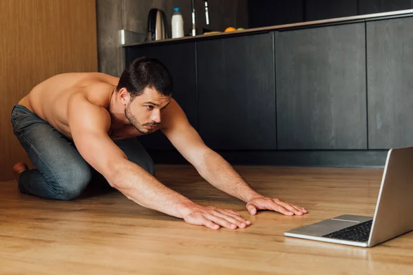 Мускулистый человек делает упражнения Balasana во время просмотра онлайн тренировки йоги на ноутбуке — стоковое фото