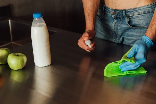 Cropped view of man holding bottle with antibacterial liquid and rag near bottle of milk and apples on wooden table — Stock Photo