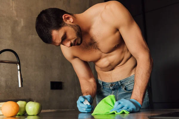 Homme musclé en gants de caoutchouc tenant chiffon et bouteille avec antiseptique près des fruits sur la table — Photo de stock