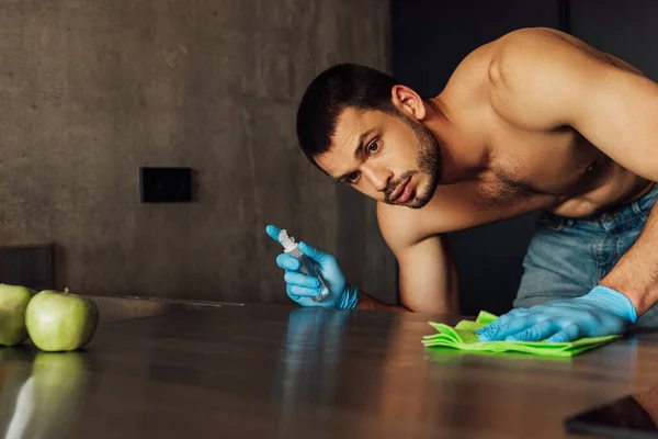 Enfoque selectivo del hombre musculoso en guantes de goma sosteniendo trapo y botella con antiséptico cerca de manzanas en la mesa - foto de stock