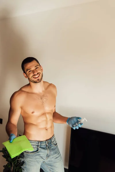 Cheerful and muscular man in rubber gloves holding rag and bottle with antibacterial liquid near tv with blank screen — Stock Photo