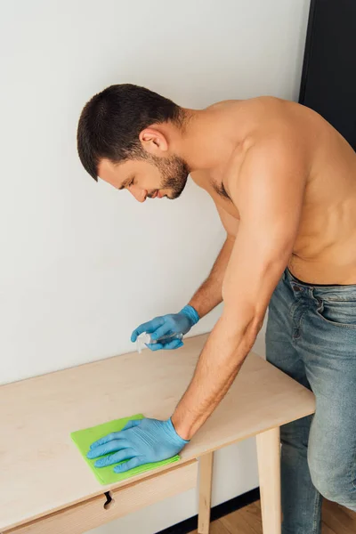 Shirtless man in rubber gloves holding rag and bottle with antibacterial liquid near table — Stock Photo