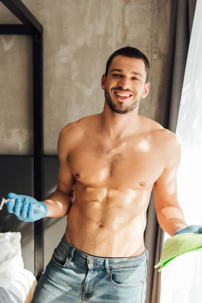 Sunlight on cheerful and muscular man in rubber gloves holding rag and bottle with antibacterial liquid at home — Stock Photo