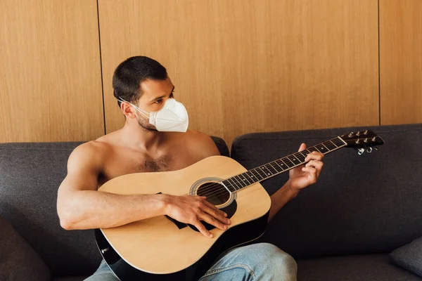 Uomo muscoloso in maschera medica suonare la chitarra acustica in soggiorno — Foto stock