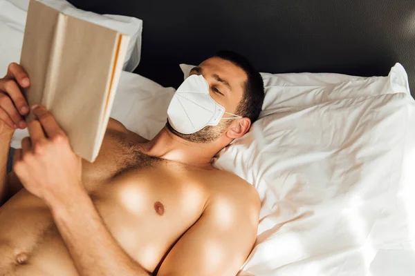 Sunlight on muscular man in medical mask reading book in bed — Stock Photo