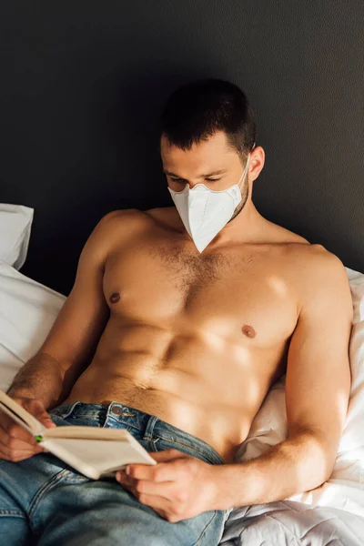 Muscular man in medical mask reading book in bed — Stock Photo