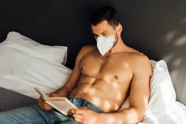 Luz del sol sobre el hombre sin camisa en la máscara médica libro de lectura en la cama - foto de stock