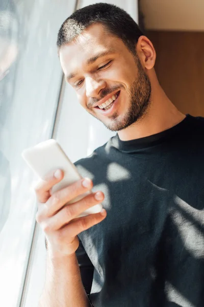 Vista de ángulo bajo del hombre feliz utilizando el teléfono inteligente cerca de la ventana en casa - foto de stock