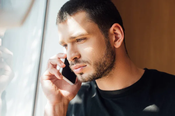 Hombre barbudo hablando en el teléfono inteligente cerca de la ventana en casa - foto de stock