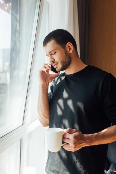 Homme barbu tenant tasse tout en parlant sur smartphone à la maison — Photo de stock