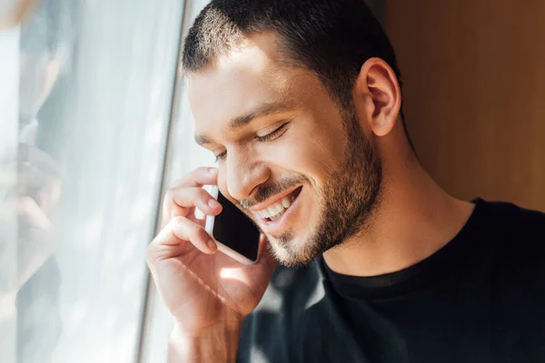 Hombre feliz hablando en el teléfono inteligente cerca de la ventana en casa - foto de stock