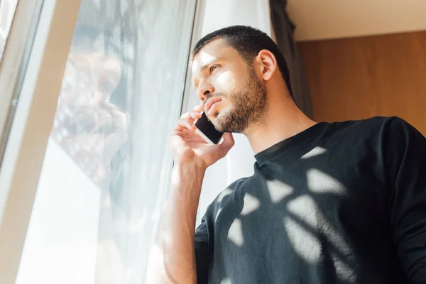 Vista a basso angolo dell'uomo che parla sullo smartphone vicino alla finestra di casa — Foto stock