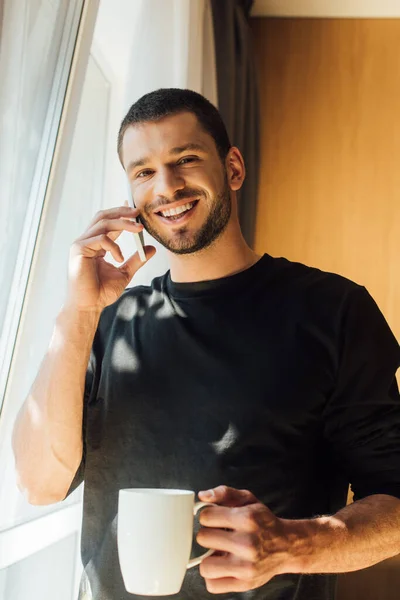 Hombre feliz sosteniendo la taza y hablando en el teléfono inteligente en casa - foto de stock