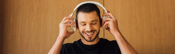 Panoramaaufnahme eines glücklichen bärtigen Mannes, der zu Hause drahtlose Kopfhörer anfasst — Stockfoto