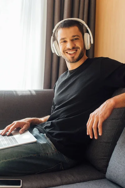 Freelancer feliz em fones de ouvido sem fio olhando para a câmera perto de laptop e smartphone com tela em branco — Fotografia de Stock