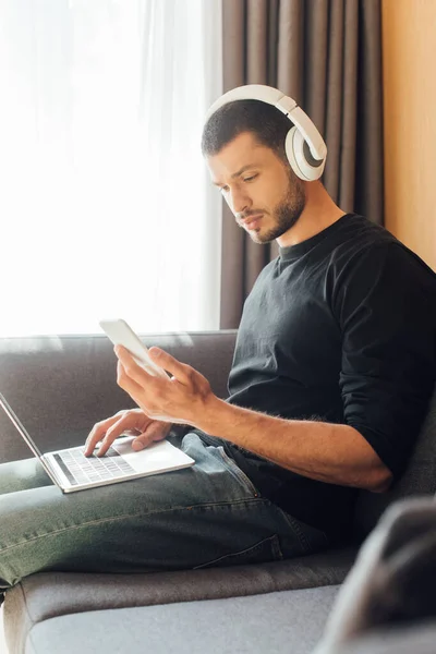 Selective focus of freelancer in wireless headphones looking at smartphone near laptop — Stock Photo