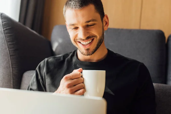 Selective focus of cheerful and bearded freelancer using laptop and holding cup — Stock Photo