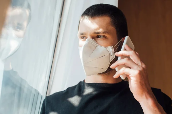 Hombre en máscara médica hablando en teléfono inteligente cerca de la ventana - foto de stock