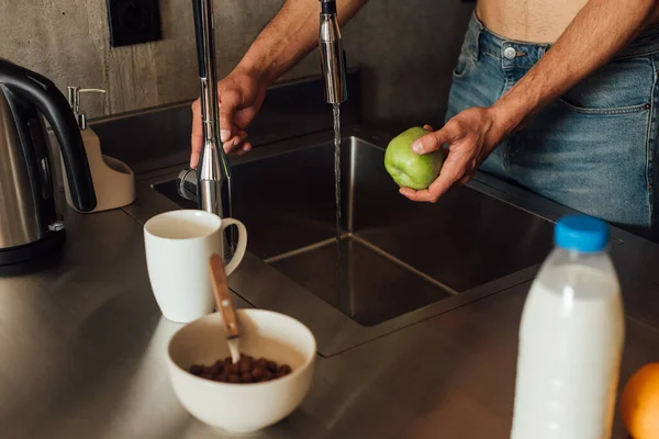 Cropped view of man holding apple near faucet with pouring water in kitchen — Stock Photo