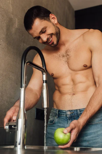 Selective focus of shirtless man smiling while holding apple with faucet with pouring water in kitchen — Stock Photo