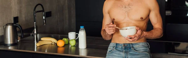 Concetto panoramico di uomo senza camicia che tiene ciotola con cornflakes vicino a frutta e bottiglia con latte in cucina — Foto stock