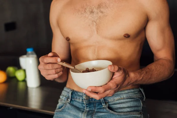 Vista recortada del hombre sin camisa sosteniendo tazón con copos de maíz cerca de frutas y botella con leche en la cocina - foto de stock